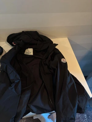Svart vindjacka från Moncler - Snygg svart vindjacka från Moncler med huva och dragkedja. Jackan har en vit och röd rand över ryggen samt Moncler-logga på ärmen. Tillverkad i lätt och tåligt material, perfekt för blåsiga dagar. Klassisk och sportig design.
