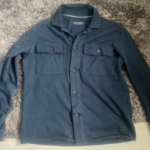 Mörkblå overshirt från Marc O'Polo - Stilren mörkblå overshirt från Marc O'Polo med klassisk krage, knäppning framtill och två bröstfickor med lock. Tillverkad i mjukt tyg som ger en avslappnad look. Storlek M