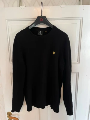 Svart stickad tröja från Lyle & Scott - Snygg svart stickad tröja från Lyle & Scott med rund halsringning och den klassiska gula logon broderad på bröstet. Tröjan har långa ärmar och är perfekt för lager på lager. Enkel och stilren design som passar till det mesta.