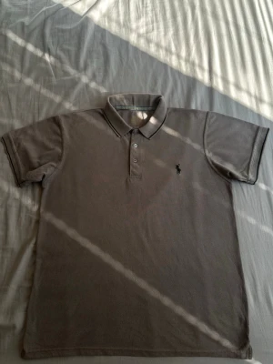 Beige pikétröja från Polo Ralph Lauren - Klassisk beige pikétröja från Polo Ralph Lauren med svart broderad logga på bröstet. Tröjan har krage, knäppning med tre knappar och korta ärmar. Perfekt till en avslappnad men ändå stilren look.