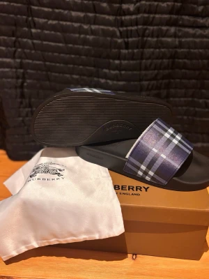 Burberry tofflor  - Säljer ett par snygga slides från Burberry med klassiskt blått och vitt rutmönster på remmen. Sandalerna har svart sula i gummi och en bred rem i textil med Burberrys ikoniska design. Perfekta för sommaren och riktigt trendiga.
