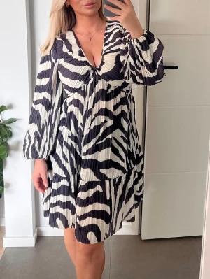 Zebramönstrad v-ringad klänning -  zebramönstrad klänning med v-ringning och långa, vida ärmar. Klänningen har plisserat tyg och en loose passform som ger en avslappnad och trendig look. Perfekt för dig som vill sticka ut med ett djurmönster.