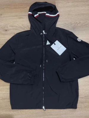 Moncler windbreaker  - Snygg svart vindjacka från Moncler med huva och dragsko. Jackan har hel dragkedja framtill, elastiska muddar och Moncler-logga på ärmen. Huvan har en vit och röd rand som detalj. Materialet är tunt och vindtätt, perfekt för blåsiga dagar.