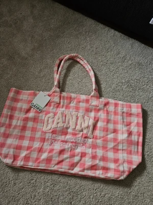 Rosa rutad Tote Bag från GANNI - Säljer en rosa och vit rutad shoppingväska från GANNI med broderad logga och texten 'Have a nice day! Please recycle' på framsidan. Väskan har dubbla handtag och är gjord i ett mjukt tygmaterial. Perfekt för att bära med sig allt man behöver.
