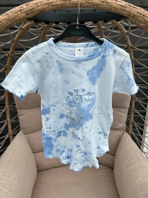 Tie dye t-shirt strl. 140 - Säljer en ljusblå batik t-shirt med korta ärmar och broderat fjärilsmotiv framtill. T-shirten har vågig kant nertill och är gjord i mjuk bomull. Perfekt för dig som gillar unika detaljer och sköna material.