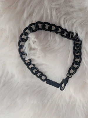 Svart kedjearmband från ICON - Snyggt svart armband från ICON med chunky kedjelänkar i matt finish. Armbandet har en robust karbinhake och en liten bricka med märkesnamnet ingraverat. Perfekt accessoar för en edgy och modern stil.