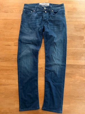 Jacob Cohën jeans - Snygga blå jeans från Jacob Cohen i klassisk rak modell. Jeansen har knäppning med silverfärgade knappar och fem fickor. Materialet är mjukt denim med lätt tvättad look och diskreta slitningar. Perfekta för en avslappnad och trendig stil. Midja: 40 cm. Längd fram: 24 cm. Total längd: 104 cm. Innersöm från grenen: 80 cm. Innersöm nertill: 18 cm.