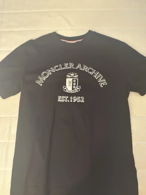 Svart Moncler Archive t-shirt - Snygg svart t-shirt från Moncler med trycket 'Moncler Archive EST. 1952' och ett emblem på bröstet. Klassisk rund hals och korta ärmar. Tillverkad i mjuk bomull och perfekt för en clean och stilren look.