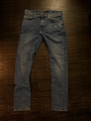 Ljusblå jeans från Nudie  - Säljer ett par ljusblå jeans från Nudie  med smal passform och klassisk femficksdesign. Jeansen har slitna detaljer och är tillverkade i denim med synliga sömmar. Perfekta för en avslappnad och trendig stil.