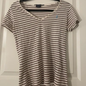 Randig t-shirt från Ralph Lauren Sport - Säljer en superfin randig t-shirt från Ralph Lauren Sport. Den är vit med bruna ränder och har en liten blå logga på bröstet. T-shirten är i 100% bomull och har en v-ringad hals. Perfekt för en avslappnad stil!💖💖💖