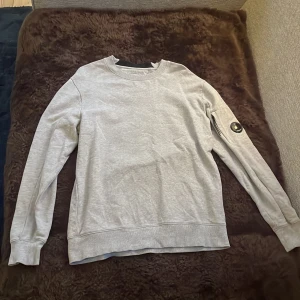 Grå tröja från C.P. Company - Säljer en stilren grå tröja från C.P. Company. Den har en cool detalj med en lins på ärmen och är perfekt för både vardag och chill. Tröjan är i bra skick och superbekväm. Perfekt för höst och vår!