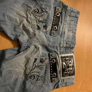 Blå jeans med broderade detaljer - Låg midja. Snygga blå jeans med unika broderade blommor på bakfickorna. Dekorerade med svarta läderdetaljer och nitar för en edgy look. Perfekta för dig som vill sticka ut!
