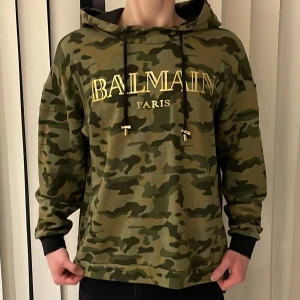 Camouflage hoodie från Balmain - Säljer en riktigt cool camouflage hoodie från Balmain med guldfärgad logga på bröstet. Tröjan har en justerbar huva med svarta snören och ribbade muddar vid ärmsluten. Skick 9/10 som ny bara utan prislapp, nypriset ligger på runt 7000kr! Perfekt för en avslappnad stil ✅