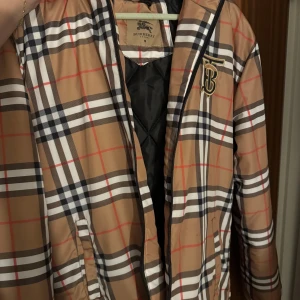 Rutig jacka från Burberry - Säljer en snygg rutig jacka från Burberry i klassiskt beige, svart, vit och röd färgkombination. Jackan har en dragkedja framtill och en broderad logga på bröstet. Perfekt för att ge din outfit en stilren touch.