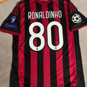Randig fotbollströja med Ronaldinho 80 - Säljer en randig fotbollströja från Adidas med Ronaldinho och nummer 80 på ryggen. Tröjan är svart och röd med korta ärmar och Champions League-märken på ärmarna. Perfekt för fotbollsfans!