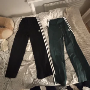 Adidas träningsbyxor med ränder - Två par Adidas träningsbyxor med klassiska vita ränder längs sidorna. Ett par är svarta och det andra är mörkgröna. Perfekta för träning eller en avslappnad stil. 