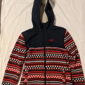 Mönstrad fleecejacka från Jack Wolfskin - Säljer en färgglad fleecejacka från Jack Wolfskin med ett geometriskt mönster i rött, svart och vitt. Jackan har en huva och dragkedja framtill. Perfekt för kyliga dagar med sin mjuka insida.