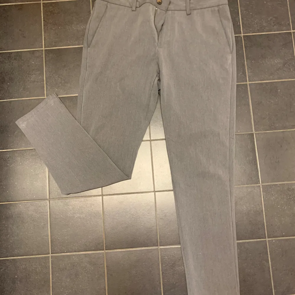 Snygga grå kostymbyxor från Lindbergh med en klassisk design. Byxorna har en dragkedja och knapp framtill samt två bakfickor med knappar. Perfekta för en stilren look. Storlek 34/32. Farkut & Housut.