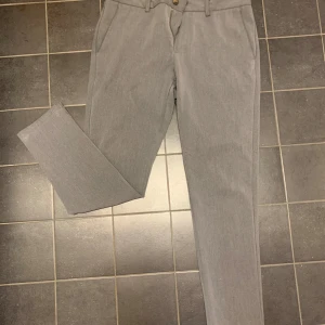 Grå kostymbyxor från Lindbergh - Snygga grå kostymbyxor från Lindbergh med en klassisk design. Byxorna har en dragkedja och knapp framtill samt två bakfickor med knappar. Perfekta för en stilren look. Storlek 34/32