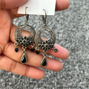 Silvriga örhängen med svarta detaljer - Eleganta silvriga örhängen med intrikata mönster och svarta pärlor. De har en rund form med hängande svarta droppar och små svarta pärlor som ger en stilfull look.