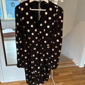 Svart klänning med prickar - Snygg svart klänning med guldiga prickar och omlott-design. Klänningen har långa ärmar och en elegant v-ringad hals. Perfekt för en festlig kväll! Ny oanvönd 