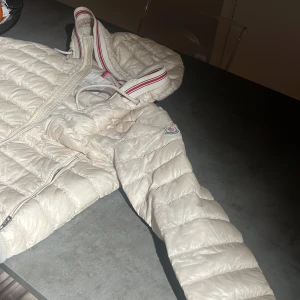 Vit pufferjacka från Moncler - Säljer en snygg vit pufferjacka från Moncler med huva och dragkedja. Jackan har ett stilrent och quiltat mönster samt Moncler-logga på ärmen. Perfekt för kyliga dagar!