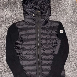 Svart dunväst med huva från Moncler - Snygg svart dunväst från Moncler med huva och dragkedja. Västen har stickade ärmar och en quiltad design. Perfekt för kyligare dagar när du vill hålla stilen. Moncler-logga på ärmen ger en exklusiv touch.