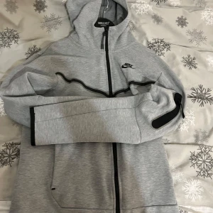 Grå hoodie från Nike - Säljer en grå hoodie från Nike med dragkedja och svart logga på bröstet. Den har en justerbar huva och långa ärmar med svarta detaljer. Perfekt för en avslappnad stil.  Pris kan diskuteras!! Slänger med byxorna till de också! 