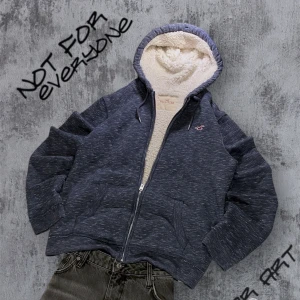 Hollister Hoodie med Foder - Säljer en mysig blå hoodie från Hollister med fleecefoder. Den har en dragkedja framtill och en broderad logga på bröstet. Perfekt för kyliga dagar med sin varma och bekväma design. Storlek L 