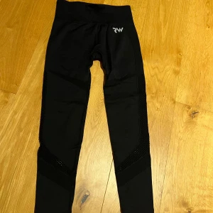 Svarta leggings från RW - Snygga svarta leggings från RW med meshdetaljer längs sidorna. Perfekta för träning eller en aktiv vardag. Tillverkade i ett stretchigt syntetmaterial för optimal komfort och rörelsefrihet.