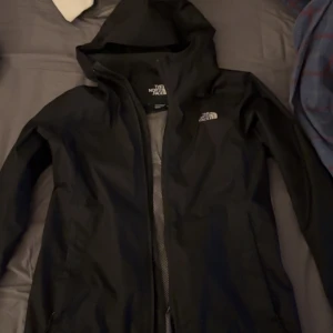 Svart vindjacka från The North Face - Säljer en svart vindjacka från The North Face i storlek XS. Jackan har en huva och dragkedja framtill, perfekt för blåsiga dagar. Den är lätt och smidig att bära med sig. Passar bra för utomhusaktiviteter och vardagsbruk.