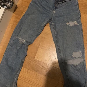 Blå jeans med slitningar - Säljer ett par blå jeans med coola slitningar på knäna. De har en klassisk femficksdesign och en knappgylf. Perfekta för en avslappnad stil.