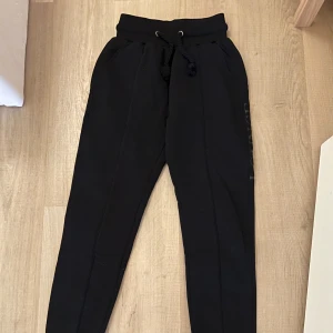 Svarta byxor och hoodie från Lounge - Säljer en svart hoodie från Lounge med dragkedja och huva. Den har långa ärmar och ett stilrent tryck längs ärmen. Säljer matchande mjukisbyxor med. Byxorna för 300, tröjan för 300 eller båda för 500, säljs ej längre och nypris var ca 1200 för båda