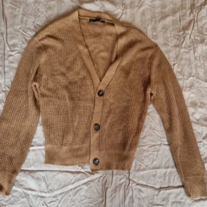 Beige ribbad kofta med knappar - Säljer en stilren beige kofta med ribbad struktur och knappar framtill. Perfekt för lager-på-lager under kyligare dagar. Koftan har långa ärmar och en v-ringad design.