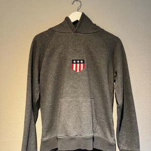 Grå hoodie från GANT - Säljer en stilren grå hoodie från GANT med broderad logga och ett emblem på bröstet. Tröjan har en klassisk känguruficka och långa ärmar. Perfekt för en avslappnad stil. Nypris 999 mitt pris endast 300. Inga tecken på andvändning. Storlek 170/15 år