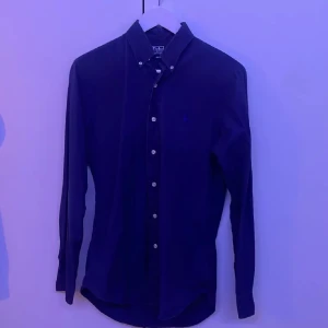 Blå skjorta från Ralph Lauren - Snygg blå skjorta från Polo Ralph Lauren med klassisk krage och knappar framtill. Skjortan har en broderad logotyp på bröstet och är i en custom fit. Perfekt för en stilren look. Nypris 1100
