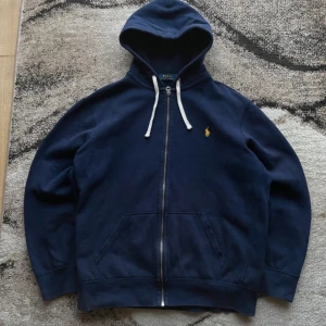Mörkblå hoodie från Polo Ralph Lauren - Säljer en snygg mörkblå hoodie från Polo Ralph Lauren med dragkedja och huva. Den har en liten gul logga på bröstet och vita snören vid huvan. Perfekt för en avslappnad stil.