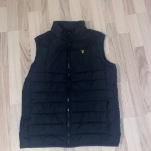 Svart dunväst från Lyle & Scott - Snygg svart dunväst från Lyle & Scott med dragkedja framtill. Västen har en quiltad design och en liten gul logga på bröstet. Perfekt för kyligare dagar.