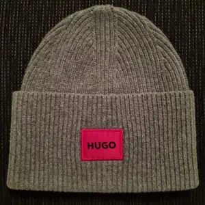 Snygg grå ribbad mössa från Hugo med en framträdande röd etikett. Perfekt för att hålla värmen under kyliga dagar. Stilren design som passar till det mesta.Helt ny oanvänd 