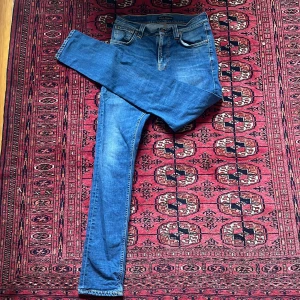 Blå jeans med broderade bakfickor - Snygga blåa nudie jeans med en klassisk femficksdesign och broderade detaljer på bakfickorna. De har en dragkedja och knapp framtill. Perfekta för en lite smalare person. Storlek 28/32