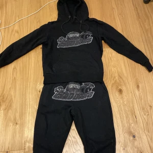 Svart hoodie från Trapstar - Säljer en svart hoodie från Trapstar med tryck på framsidan. Hoodien har en klassisk design med en stor ficka framtill och justerbar huva. Perfekt för en avslappnad stil.