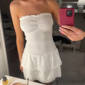 Vit axelbandslös klänning - Säljer en söt vit axelbandslös klänning med smockdetaljer och volangkjol. Perfekt för sommarens varma dagar. Klänningen har en kort längd och är tillverkad i ett lätt och luftigt material.