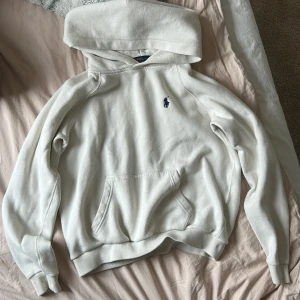 Vit hoodie från Ralph Lauren - Säljer en stilren vit hoodie från Ralph Lauren med det klassiska broderade marinblå logotypen på bröstet. Hoodien har en stor ficka framtill och en bekväm huva. Perfekt för en avslappnad look. snören saknas🫶🏼
