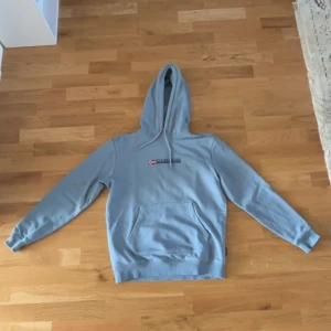 Ljusblå/Grå hoodie från Napapijri - Säljer en ljusblå hoodie från Napapijri med en klassisk logga och norsk flagga på bröstet. Tröjan har en stor ficka framtill och en bekväm huva. Perfekt för en avslappnad stil. Använd ett fåtal gånger.