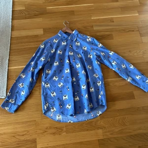Blå skjorta från ralph lauren  - Säljer en blå skjorta med ett unikt mönster av små figurer och symboler. Skjortan har långa ärmar och knappar framtill. Perfekt för dig som vill sticka ut med en färgglad och lekfull stil.
