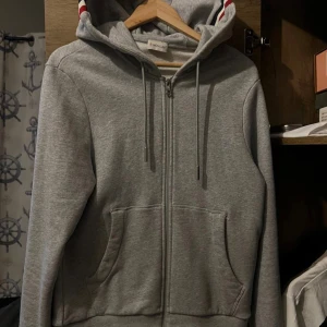 Grå hoodie från Moncler - Säljer en stilren grå hoodie från Moncler med dragkedja och justerbar huva. Tröjan har långa ärmar och en broderad Moncler-logga på ärmen. Perfekt för en avslappnad stil.