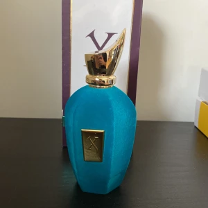 Blå parfymflaska med guldlock - Elegant blå parfymflaska från Xerjoff med ett unikt guldlock som har en böjd design. Flaskan har en rektangulär guldplatta med märkets logotyp på framsidan. Perfekt för den som söker en lyxig doftupplevelse.