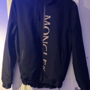 Svart jacka från Moncler - Snygg svart jacka från Moncler med dragkedja och stor logotyp på framsidan. Jackan har långa ärmar och justerbar nederkant. Perfekt för en stilren look.