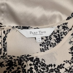 Beige och svart klänning från Part Two - Säljer en elegant beige klänning från Part Two med ett vackert svart blommönster. Klänningen har trekvartsärmar och en lös passform, perfekt för både vardag och fest. Den kommer med ett matchande underklänning i beige. Materialet känns mjukt och lyxigt.