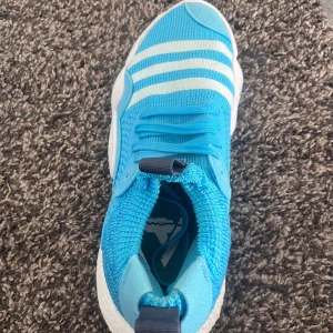 Blå sneakers från Adidas - Säljer ett par snygga blå sneakers från Adidas. De har en stickad ovandel med vita ränder och en bekväm vit sula. Perfekta för både träning och vardagsbruk. Skorna har snörning och en modern design.